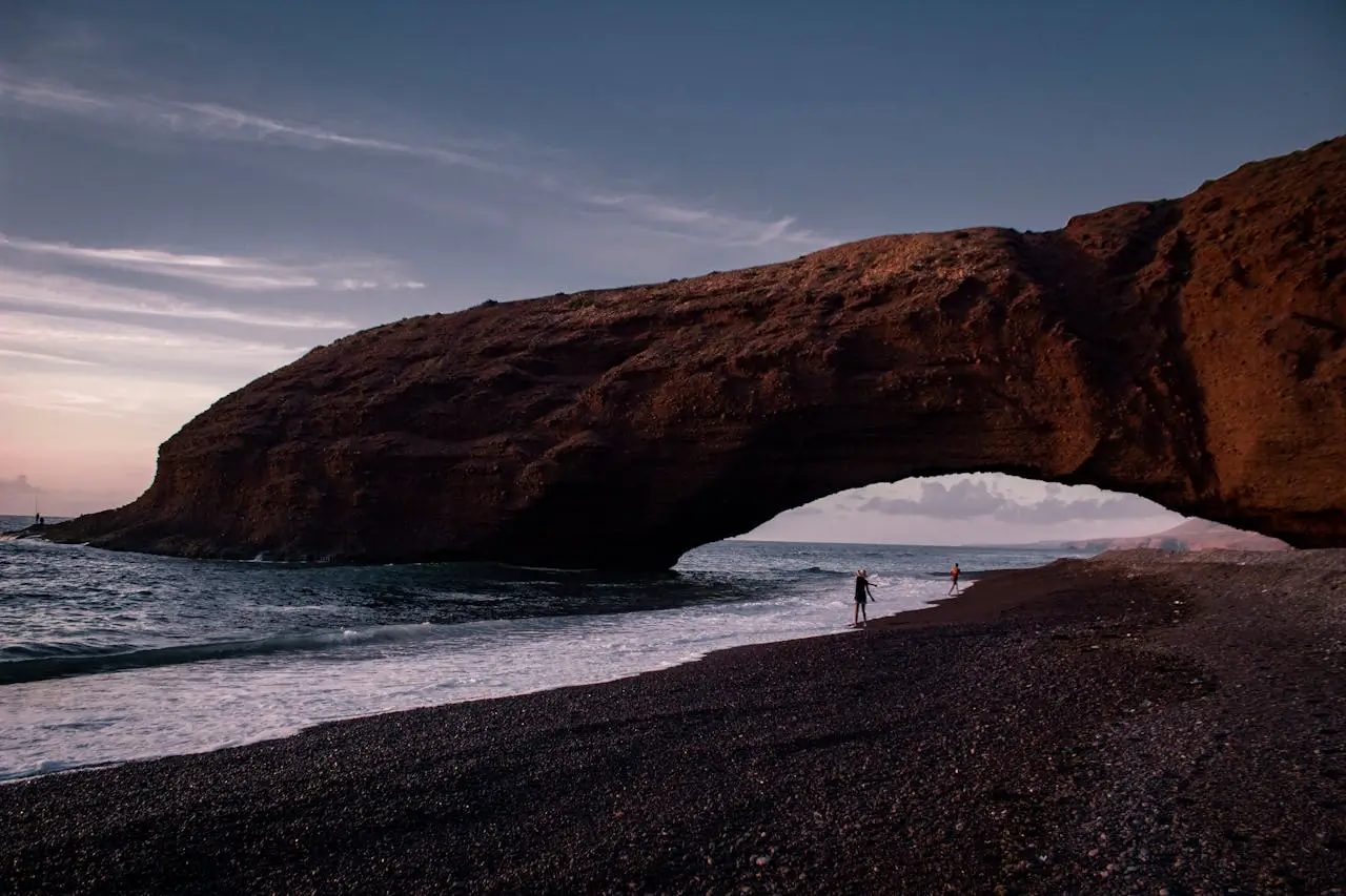 Natural rock arch on a black sand beach along the Atlantic coast of Morocco with people walking by the sea at sunset