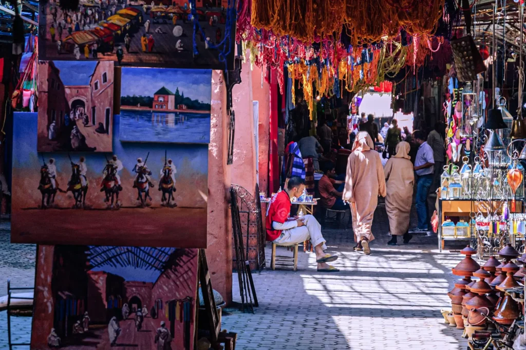 Colorful Moroccan souk in Marrakech Medina with handmade crafts, traditional paintings, clay tagines, and local people walking through the market.