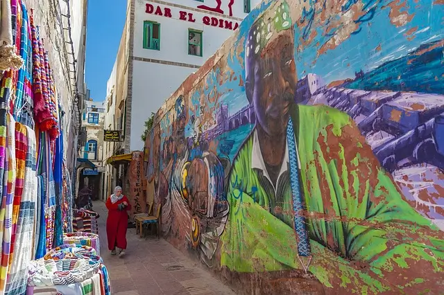 A colorful street in the medina of Essaouira, Morocco, featuring hanging textiles, a woman in a red djellaba walking past a large, textured mural of a man.