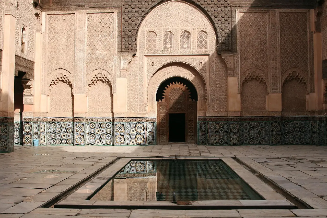 Courtyard of a Moroccan building with a central reflecting pool, intricate carved stucco, and colorful tile mosaics.
