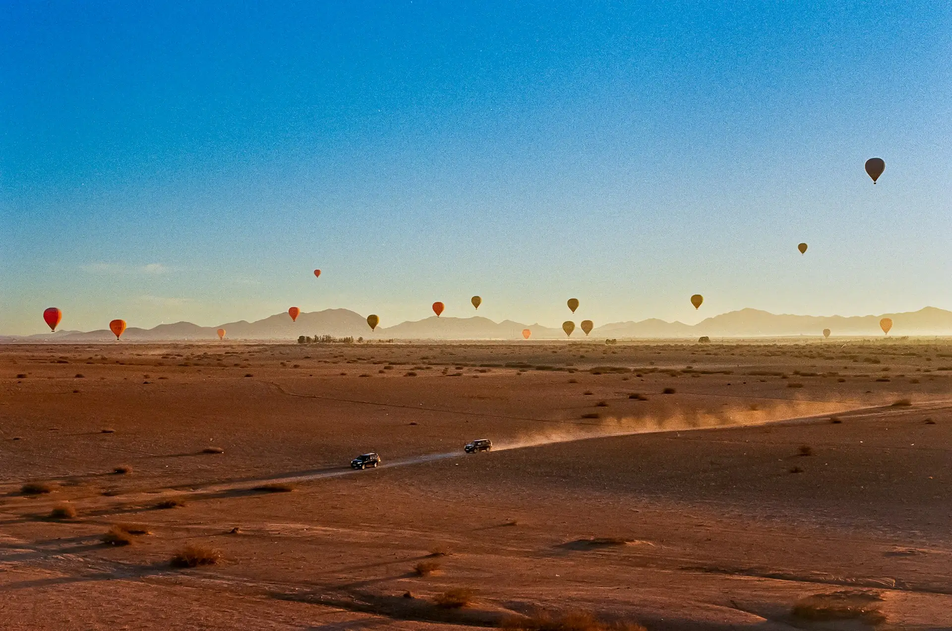 Hot air balloons floating over the Moroccan desert at sunrise with 4x4 vehicles driving across the landscape