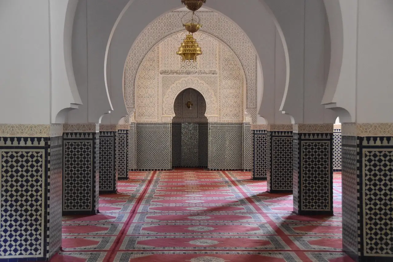 Interior of a Moroccan mosque showcasing ornate arches, geometric tilework (zellige), and a richly patterned carpet.