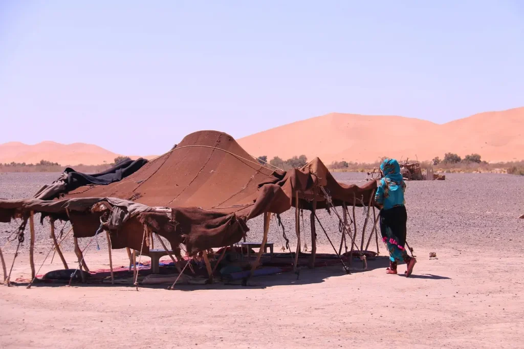 A traditional Berber tent set up in the arid Moroccan desert, with a woman in colorful attire standing nearby under a clear sky.