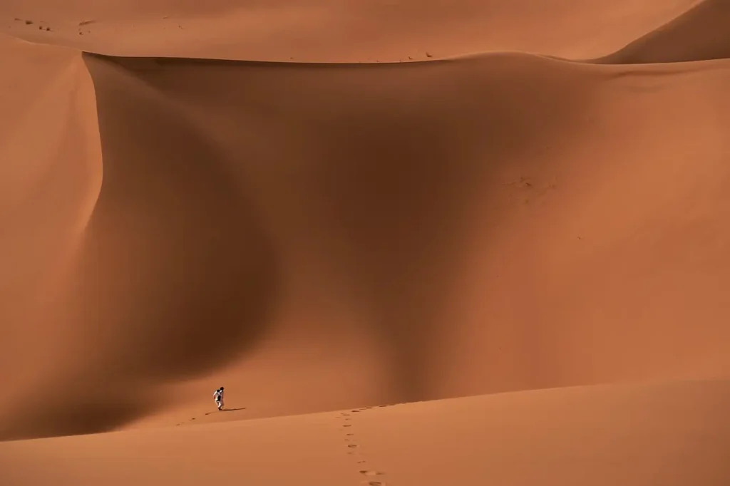 A solitary figure walking across vast, undulating desert sand dunes in Morocco, leaving footprints in the golden sand.