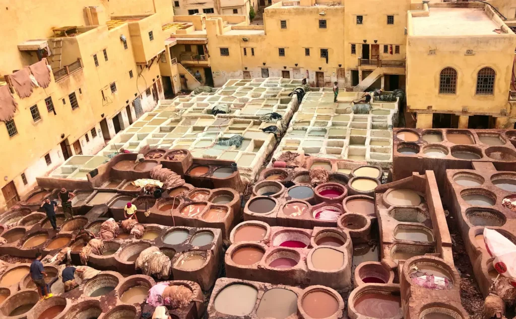 An aerial view of the ancient tanneries in Fes, Morocco, with numerous stone vats filled with colorful dyes and workers processing leather.