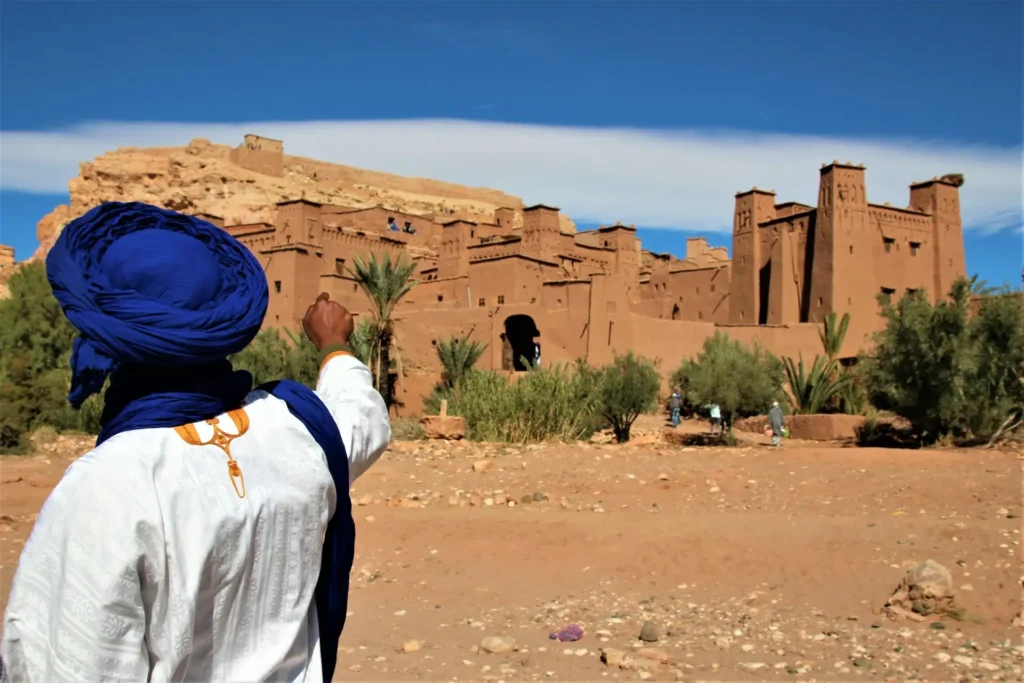 A man in a blue turban looks towards the ancient clay kasbah of Ait Benhaddou in Morocco under a bright blue sky.